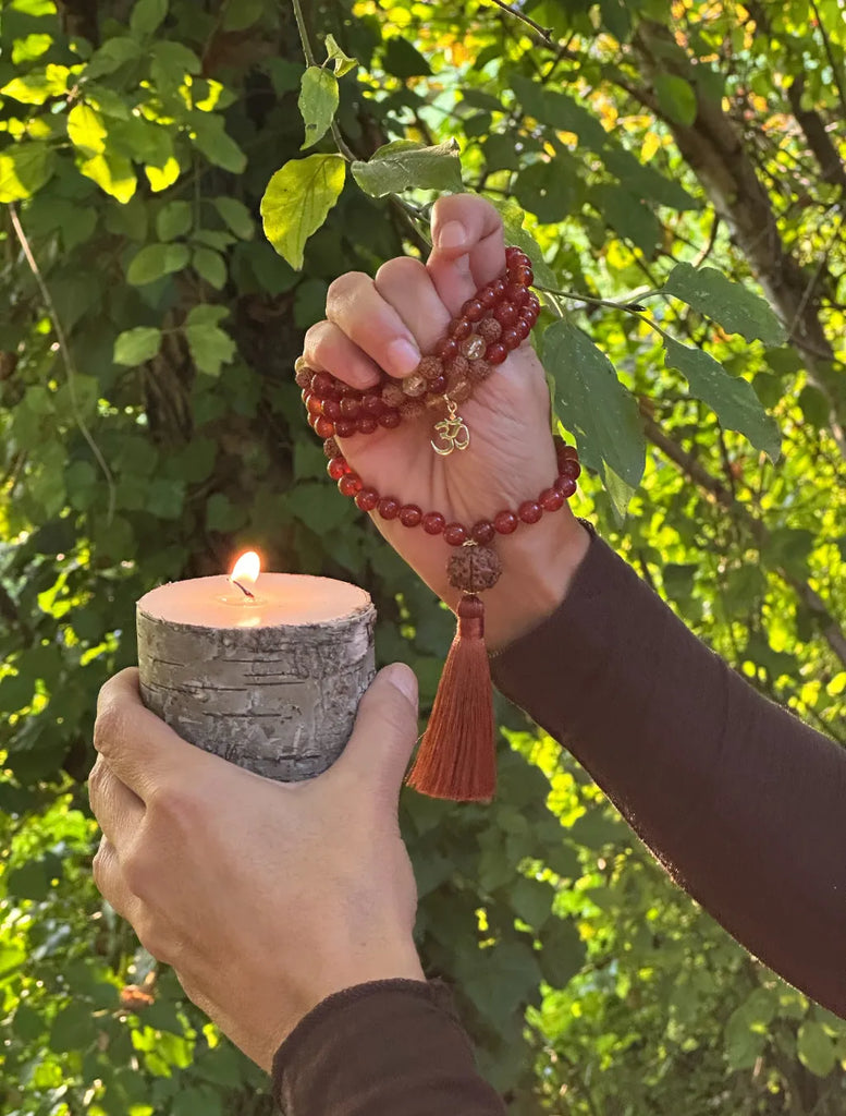 Japamālā Diwali in Rudrākṣa, Corniola e Quarzo Citrino tenuta in mano in posizione di mudra, con una candela accesa sotto a evocare la luce di Dīpāvalī.