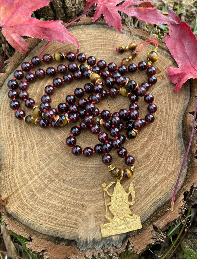 Japamālā Kalī in Granato, Ematite e Occhio di Tigre distesa per intero, con pendente Kalī in argento dorato e dettagli in macramè visibili.