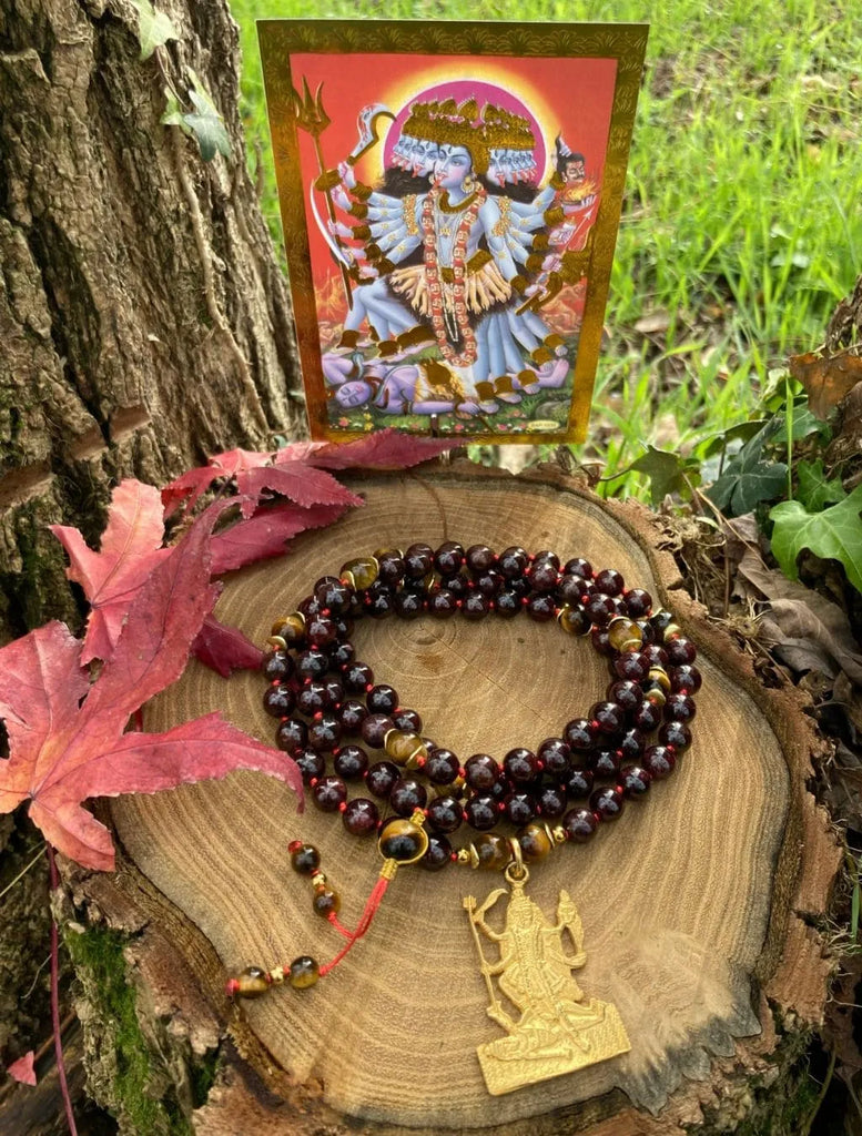 Japamālā Kalī in Granato, Ematite e Occhio di Tigre appoggiata su un tronco, con l’immagine della Dea Kalī sullo sfondo a evocare forza e trasformazione interiore.