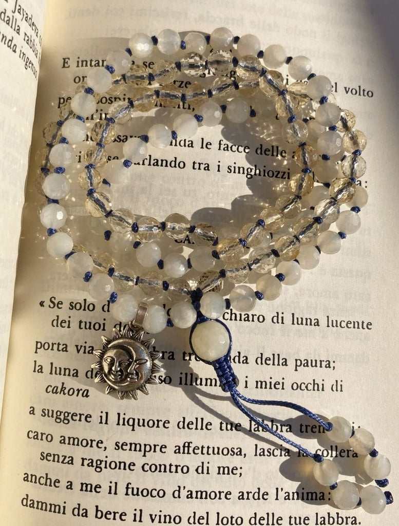 Japamala con 108+1 grani di Pietra di Luna e Quarzo Citrino, con pendente argento Surya-Chandra, arrotolata su una pagina di un libro.
